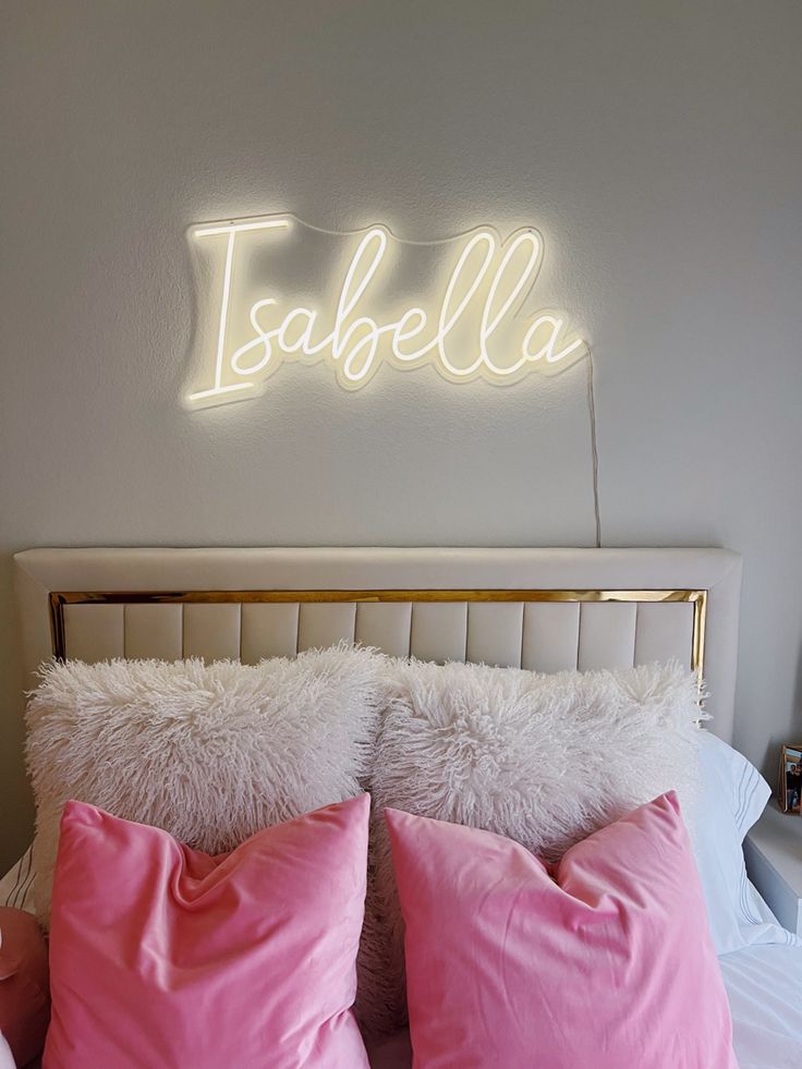 a white bed topped with pink pillows next to a neon sign above the headboard