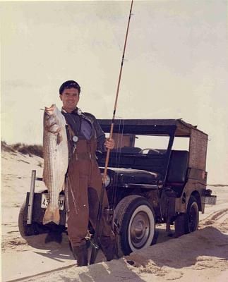 a man holding a fish while standing next to a truck on the beach with a fishing rod