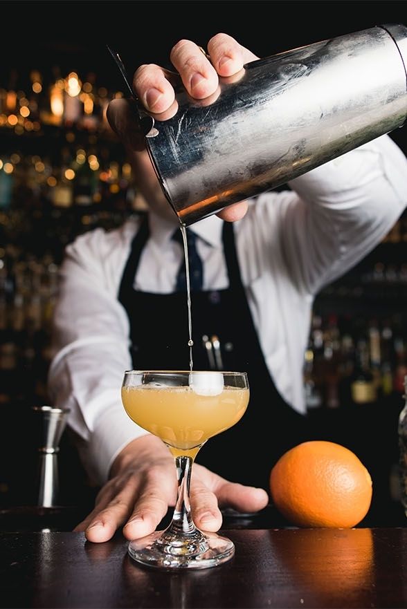 a bartender pouring a drink into a glass at a bar with an orange in the foreground