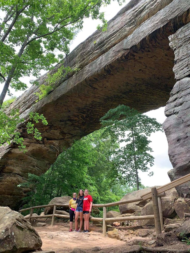 three people standing in front of a large rock arch at the top of a hill