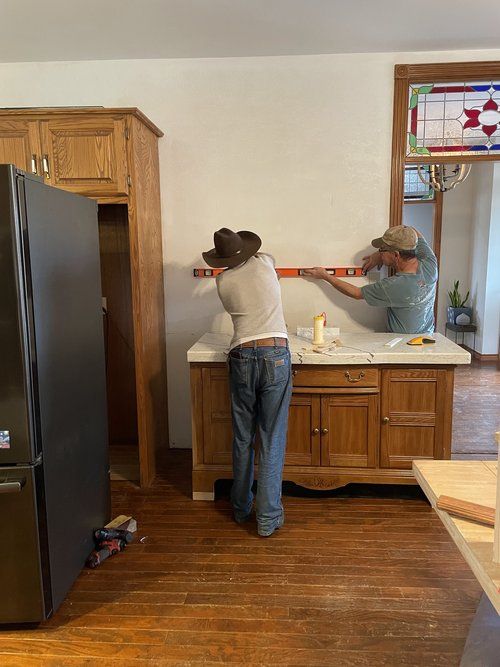 two people working on a kitchen counter in a house with wood flooring and white walls