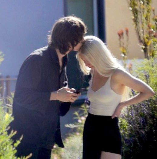 a man and woman standing next to each other in front of bushes looking at their cell phones