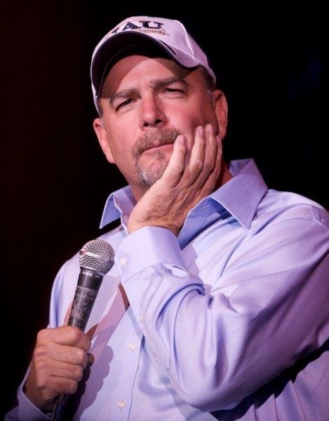 a man in a baseball cap holding a microphone and looking to the side with his hand on his chin