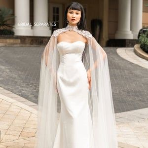 a woman wearing a white wedding gown with a long veil on her head is standing in front of a large building