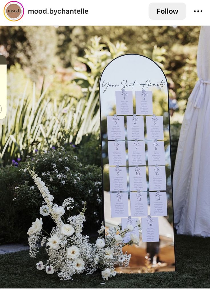 a wedding seating chart with white flowers in the foreground and an outdoor ceremony backdrop