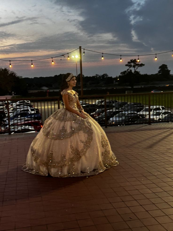 a woman in a ball gown standing on a brick walkway at night with string lights strung above her