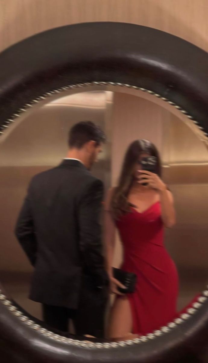 a man in a suit and woman in a red dress are looking at each other through a round mirror