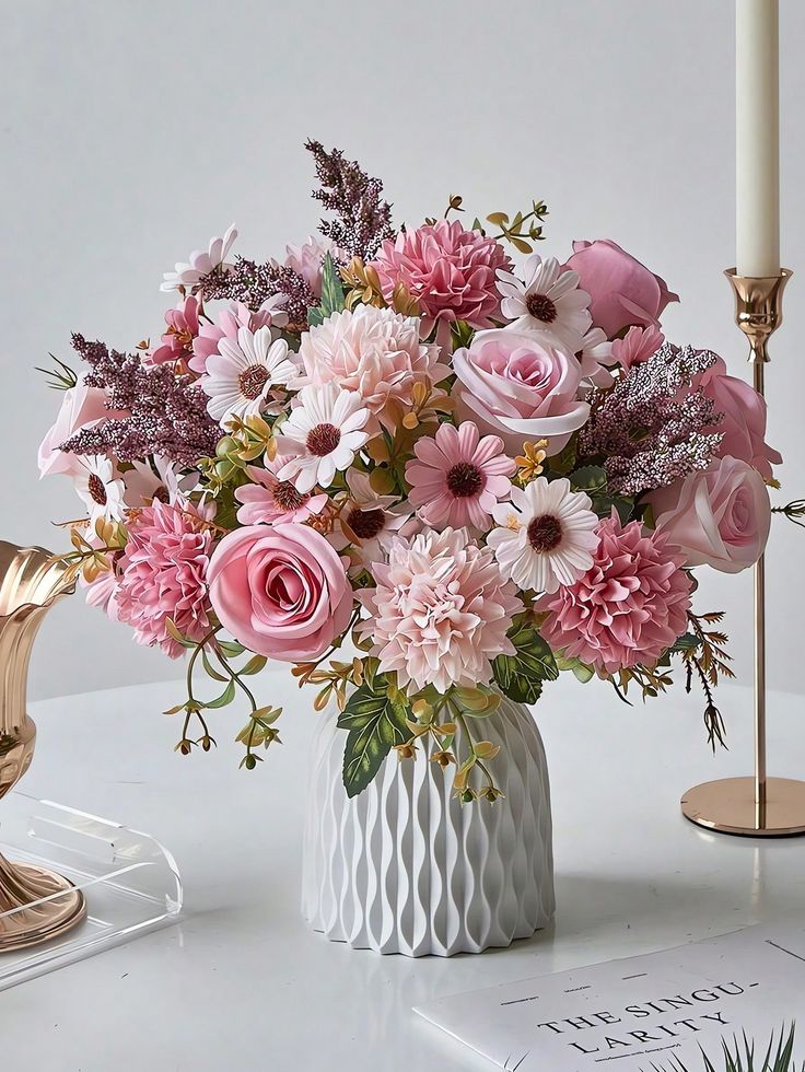 a white vase filled with pink flowers on top of a table next to a candle