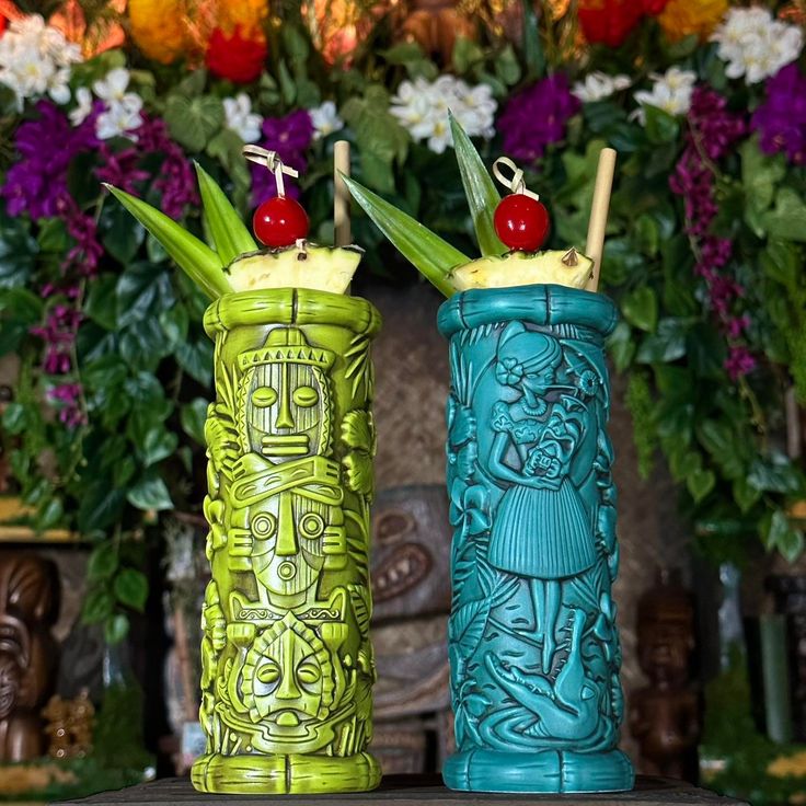 two green and blue vases sitting on top of a table with flowers in the background