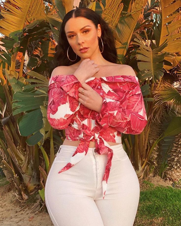 a woman wearing white pants and a pink top posing for the camera with palm trees in the background