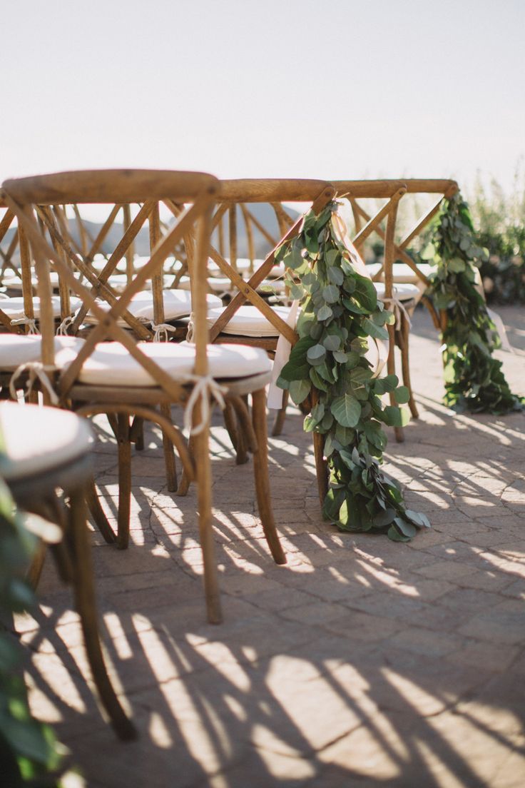 rows of wooden chairs with white cushions and greenery on the back, set up for an outdoor ceremony