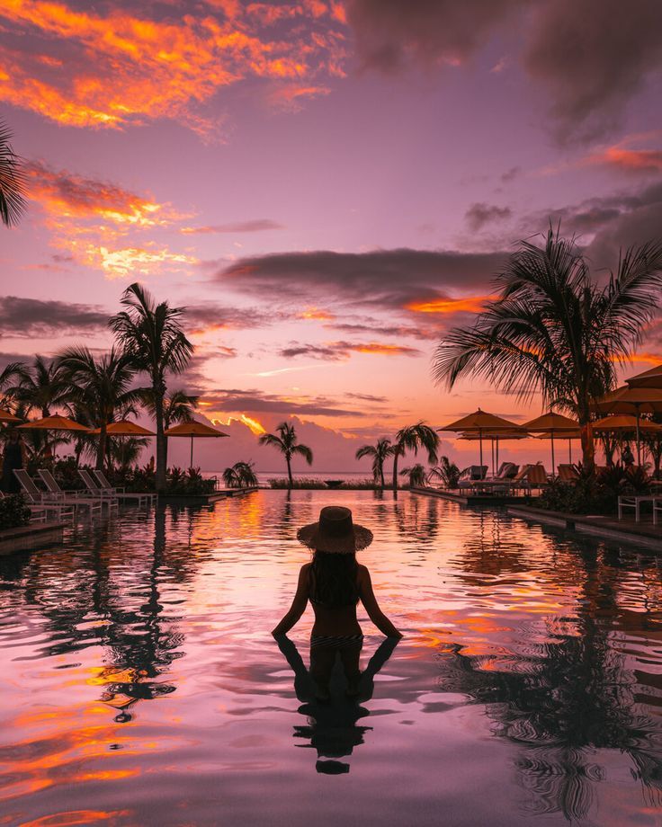 a woman standing in the middle of a swimming pool at sunset with palm trees and umbrellas