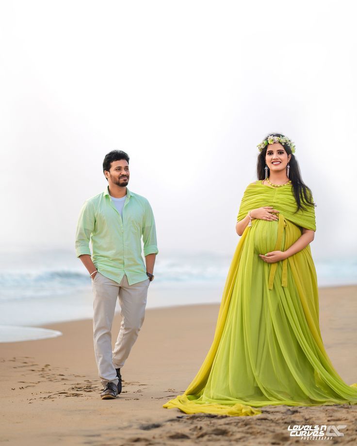 a pregnant woman in a yellow dress walking on the beach next to a man wearing a green shirt