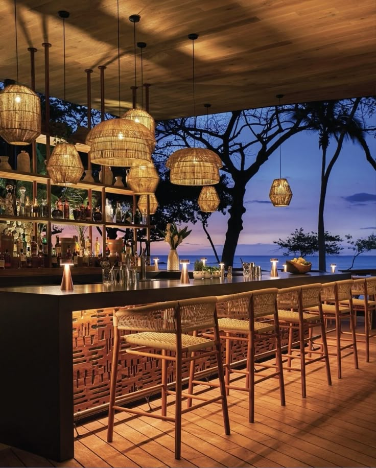 an outdoor bar with wicker chairs and hanging lights over the bar, overlooking the ocean