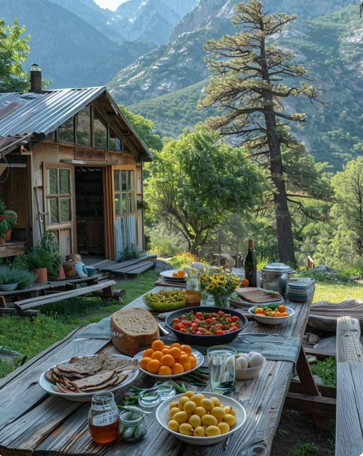 a picnic table with food and drinks on it in front of a mountain hut, surrounded by mountains