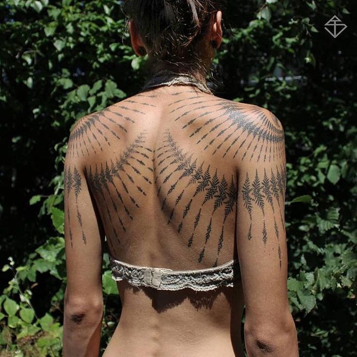 a woman with tattoos on her back standing in front of some bushes and trees,