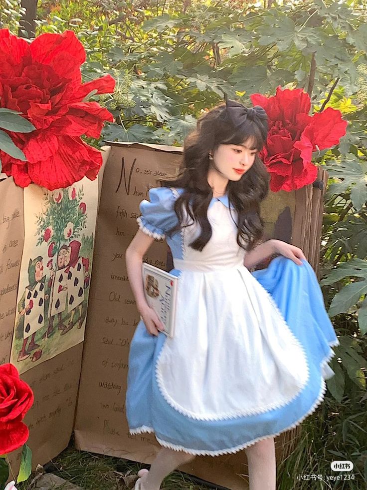 a woman in a white dress is standing next to red flowers and holding a book