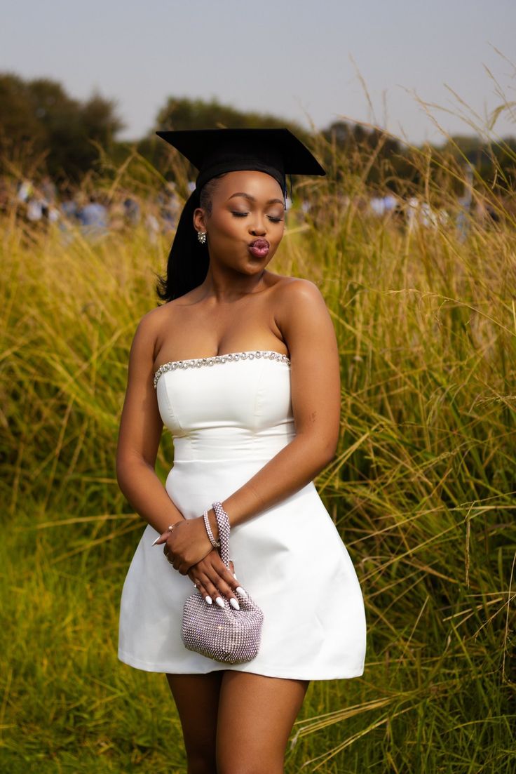 a woman wearing a graduation gown and holding a purse in front of tall grass with her hand on her hip