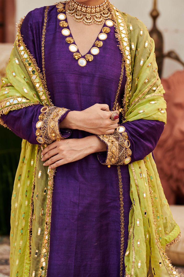 A striking ensemble, this purple raw silk sharara set showcases exquisite mirror work on the neckline and sleeves of the tunic, exuding elegance and charm. Paired with matching pants and a contrasting lime green organza dupatta, this outfit beautifully balances richness and vibrancy, making it a perfect choice for festive celebrations and special events. Anarkali Purple Palazzo Set With Gota Work, Designer Purple Kurta With Gota Work, Designer Purple Chanderi Palazzo Set, Purple Palazzo Set With Mirror Work For Festive Occasions, Purple Slub Silk Wedding Set, Purple Anarkali Palazzo Set In Chanderi, Festive Purple Kurta With Gota Work, Festive Purple Palazzo Set With Dupatta, Purple Anarkali Chanderi Palazzo Set