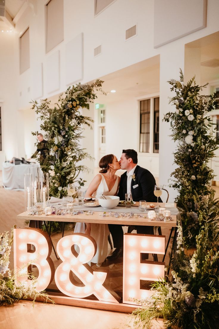 a bride and groom kissing in front of a light up b & e sign at their reception