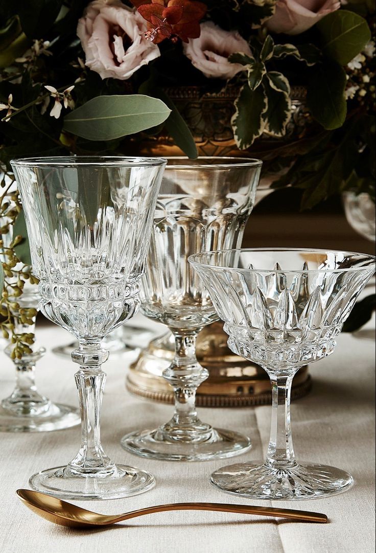 an assortment of crystal goblets on a table with flowers in the background