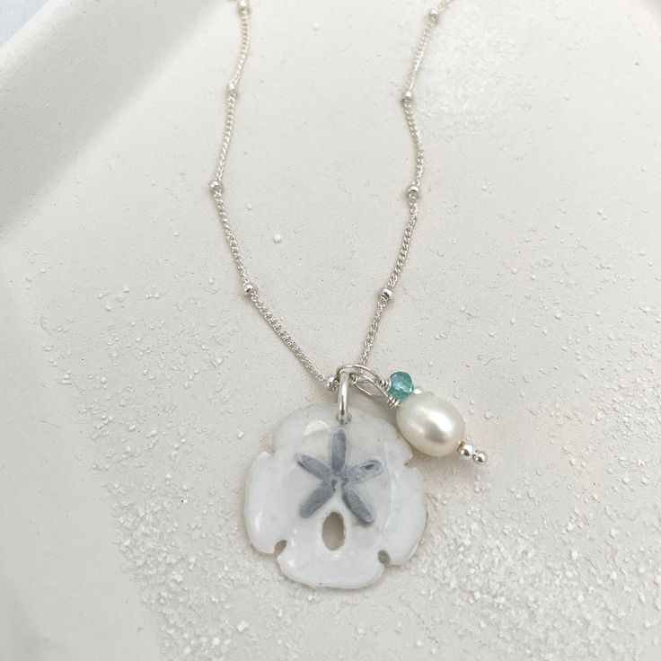 Legend has it that finding a sand dollar during a beach stroll is considered a lucky omen. So take a little bit of the beach and good luck with you wherever you go with our gorgeous Sand Dollar Charm Necklace. Features our most loved satellite chain, freshwater pearl, and blue zircon accents. Freshwater Baroque Pearl size: 6 to 8mm Blue Zircon roundel gemstone 4mm 14kt gold fill or sterling silver round beads 2mm 14kt gold fill or sterling silver satellite chain Real sand dollars are preserved a Gift Ocean-inspired Shell Necklace With Starfish Charm, Silver Necklace With Pearl Charm For Beach, White Ocean-inspired Charm Necklace For Gift, Coastal Style Shell Strand Necklace As Gift, Ocean-inspired White Charm Necklace As Gift, Ocean-inspired White Charm Necklace Gift, Coastal Style Strand Shell Necklace As Gift, Ocean-inspired Strand Charm Necklaces As Gift, White Starfish Shell Necklace As A Gift