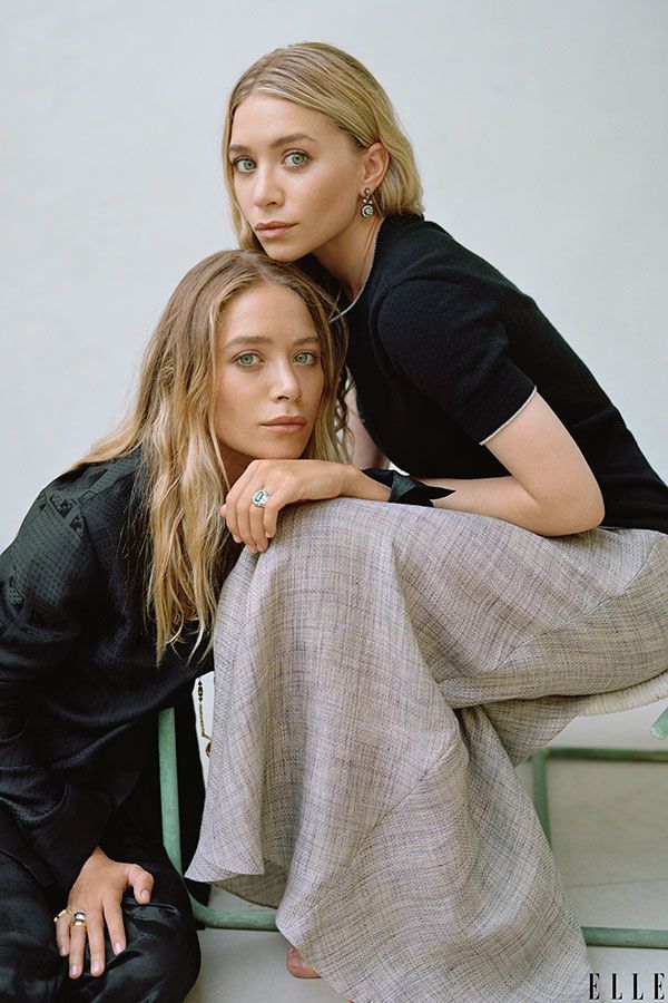 two beautiful women sitting next to each other in front of a white wall and chair