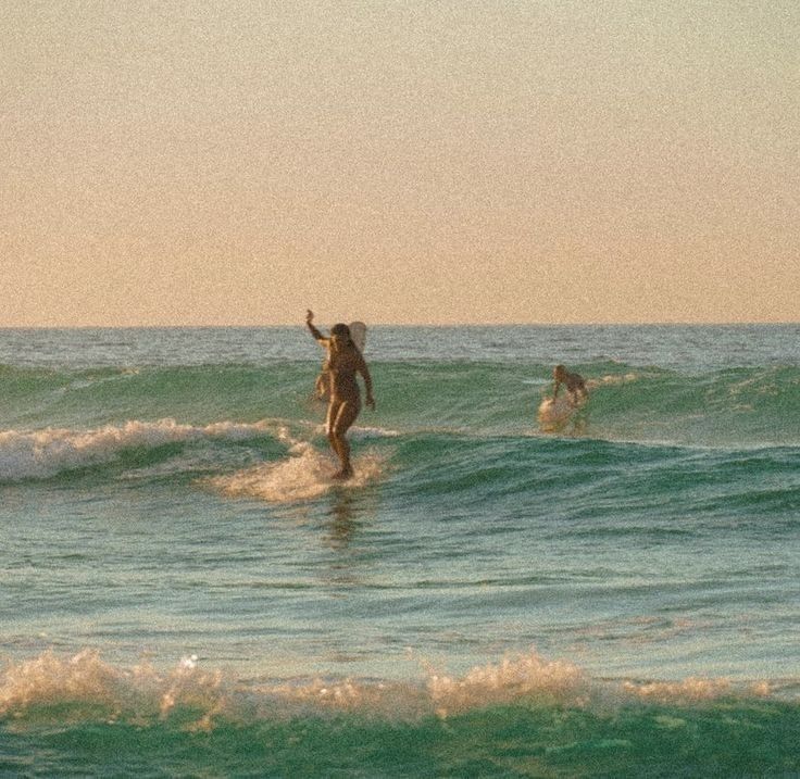 two people in the ocean on surfboards with one holding up her hand and another standing at the edge of the water