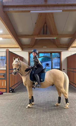 a woman riding on the back of a brown horse in an indoor arena with wooden stalls