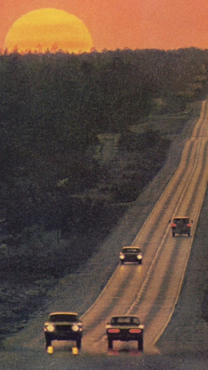 cars driving down the road in front of the sun at sunset or sunrise, with trees and bushes on either side