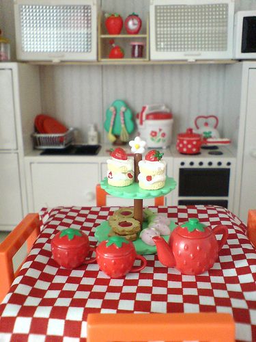 a doll house kitchen with red and white checkered table cloth, orange chairs, and toy food