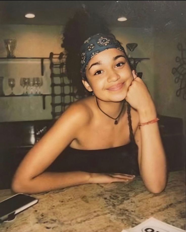 a young woman sitting at a table with her hand on her chin and looking into the camera