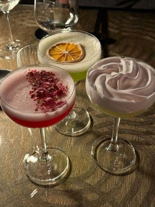 three glasses filled with different types of drinks on top of a wooden table next to wine glasses