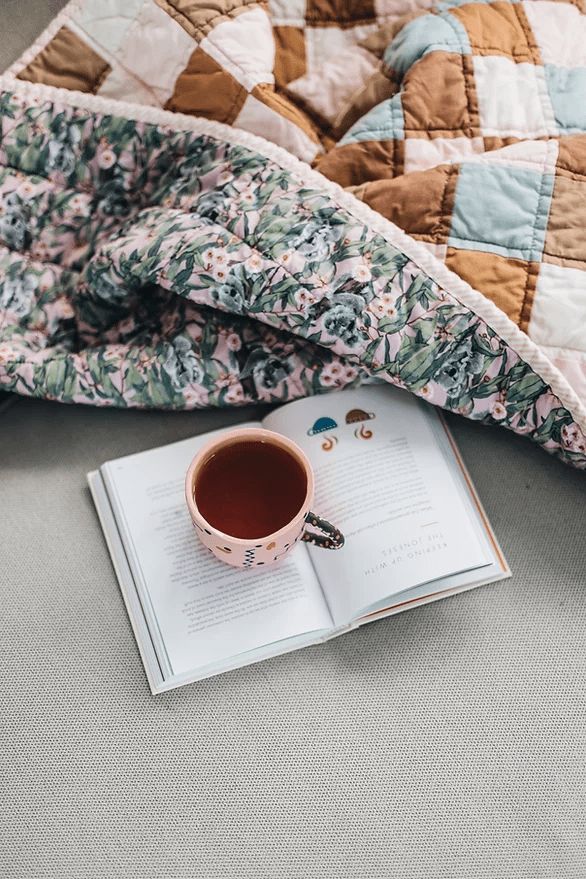 an open book with a cup of tea on top of it next to a quilt