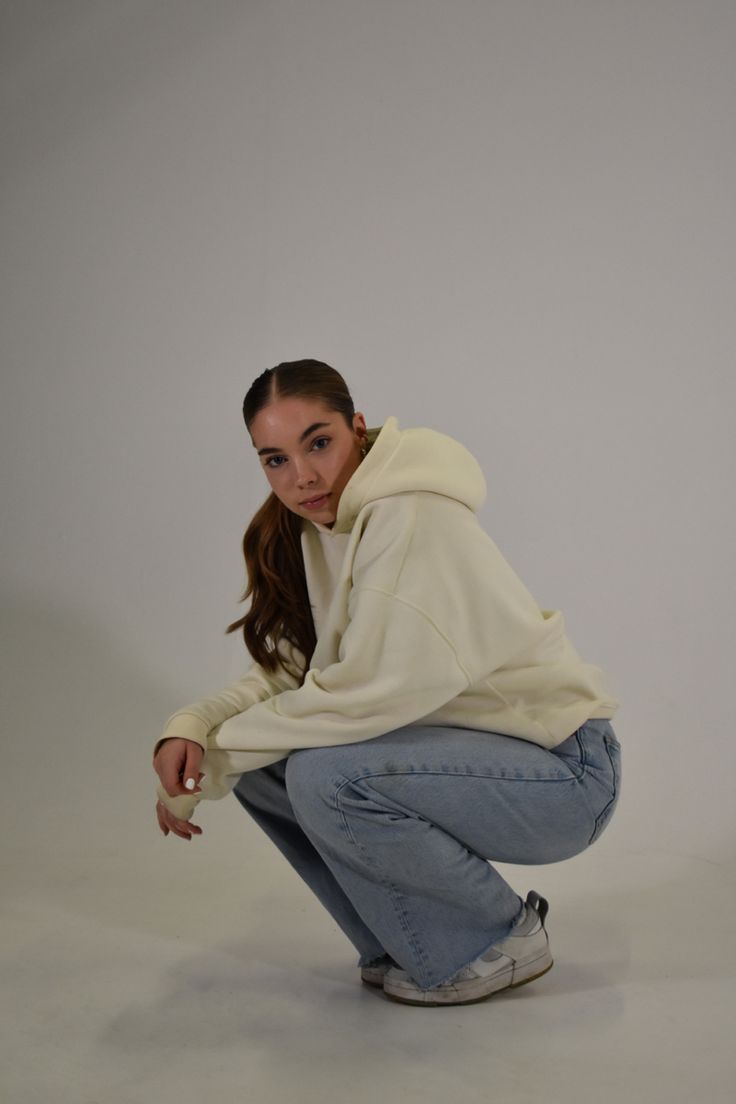 a woman squatting down in front of a white background wearing jeans and a hoodie