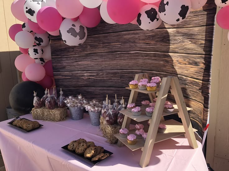 a table topped with lots of desserts next to a wooden wall covered in pink and white balloons