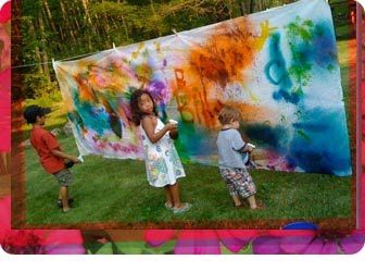 two children are painting on a large piece of art in the grass while another child is standing next to them