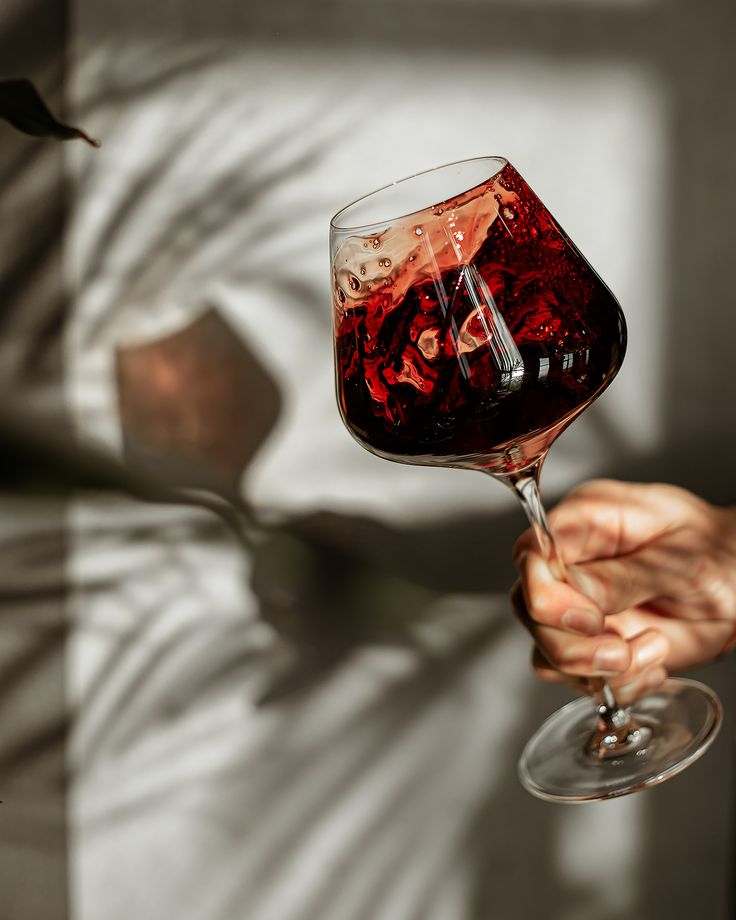 a person holding a wine glass with red liquid in it and the shadow of a tree behind them