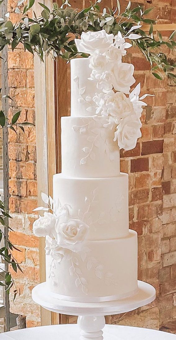 a three tiered white wedding cake with flowers on top and greenery around it