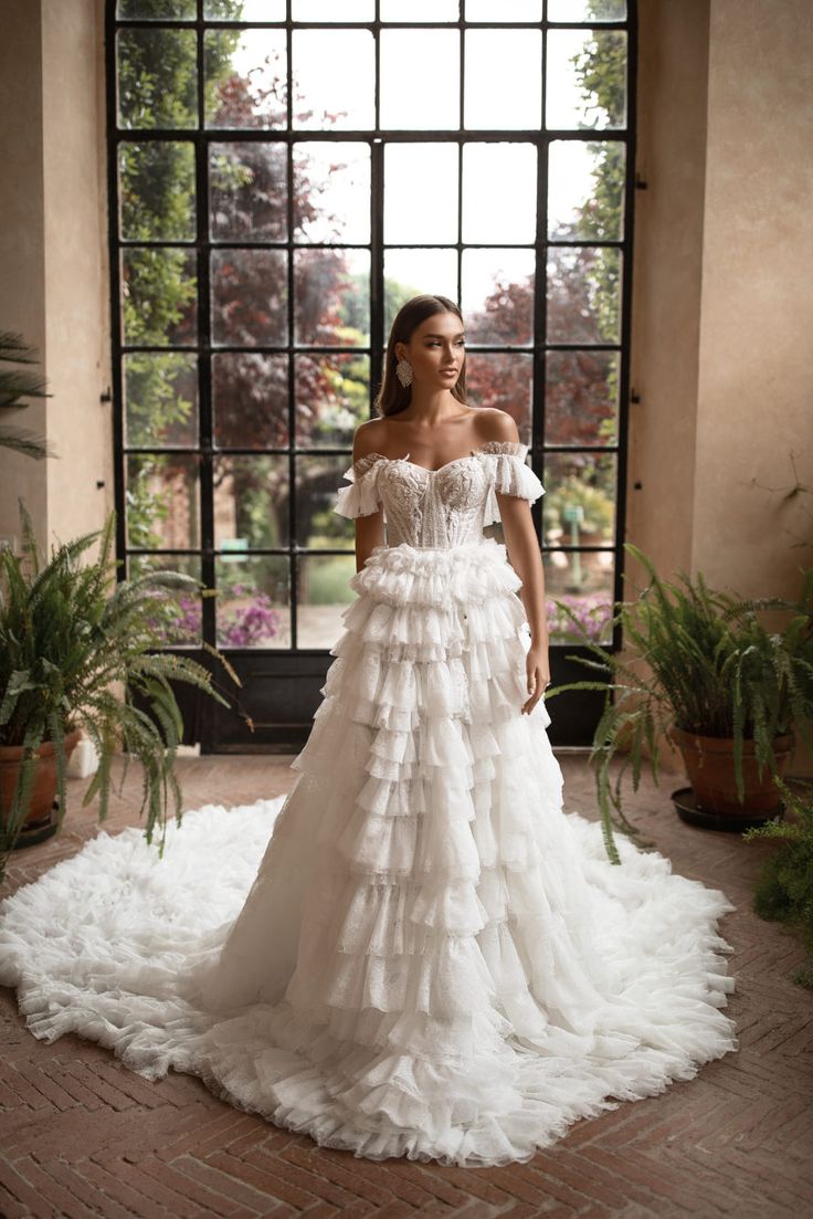 a woman standing in front of a window wearing a wedding dress with ruffled layers