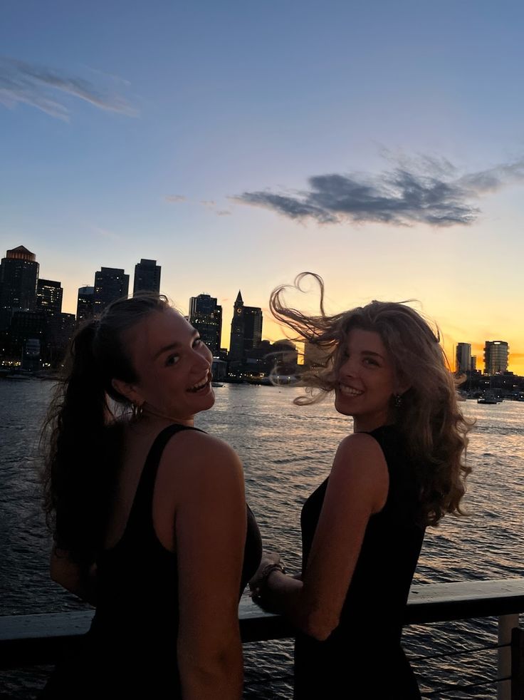 two women standing next to each other in front of a body of water at sunset