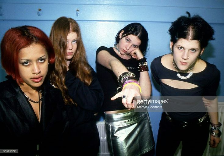 four young women posing in front of a blue wall with one pointing at the camera