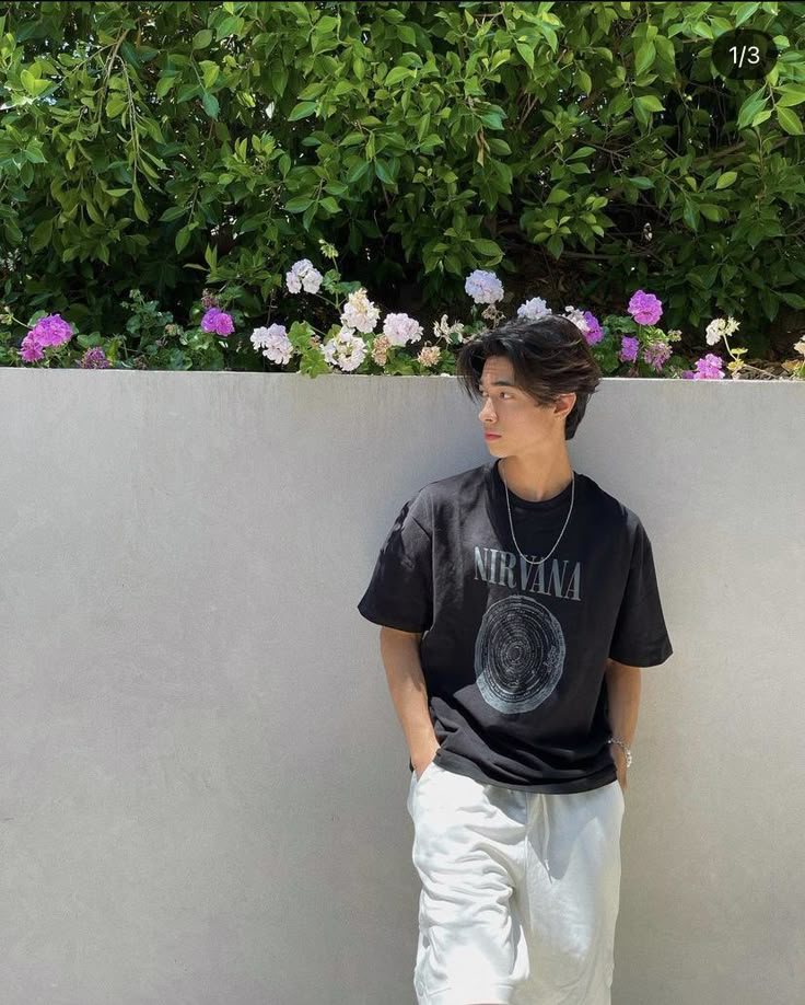 a young man leaning against a wall with his hands in his pockets, wearing white pants and a black t - shirt