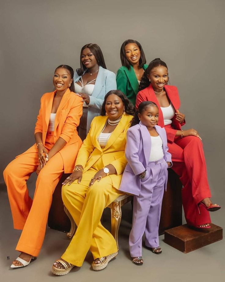 the women are posing for a photo together in bright colored suits and matching shoes, with one woman sitting on a chair