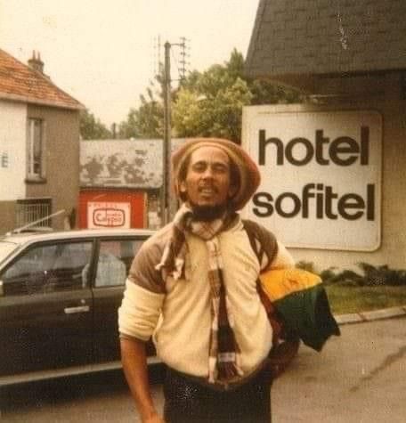 a man standing in front of a hotel with a hat on his head and scarf around his neck