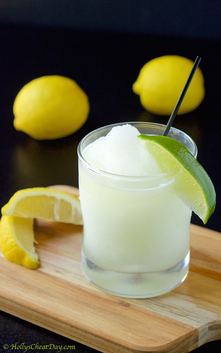 a glass filled with ice and lime on top of a wooden cutting board next to lemons