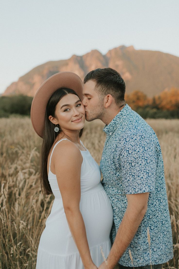 a pregnant couple kissing in the middle of a field with mountains in the back ground