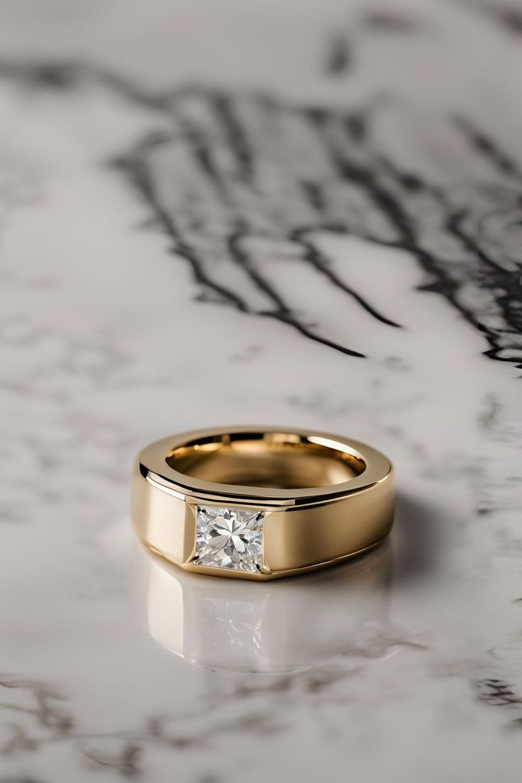 a gold wedding ring with a princess cut diamond on the side, sitting on a marble surface