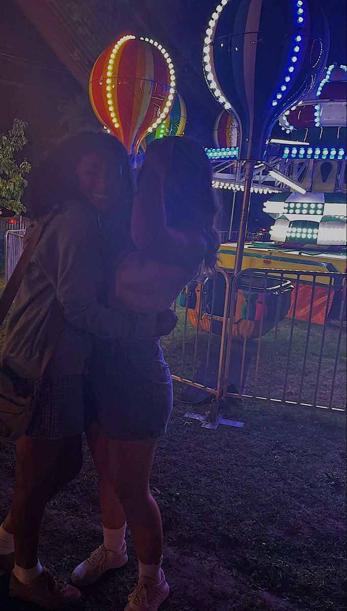 two women hugging each other in front of an amusement park ride at night with lights on