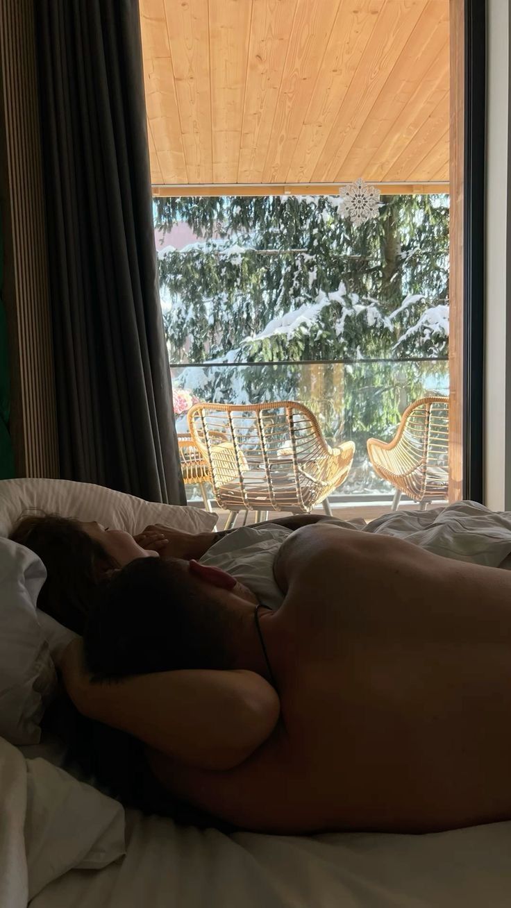 a naked man laying on top of a bed in front of a window next to a chair